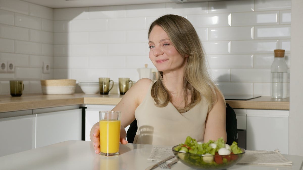 Smiling woman enjoying a healthy meal with salad and orange juice in a modern kitchen.